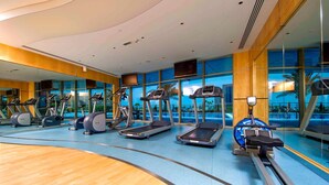 Een sportschool met loopbanden, elliptische trainers en hometrainers, grote ramen en een houten vloer.