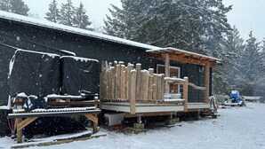 En snötäckt träkonstruktion med en terrass och ett takförsett område, omgiven av träd.