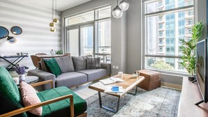 Ein modernes Wohnzimmer mit grauem Sofa, einem grünen Sessel, einem Holztisch und großen Fenstern mit Blick auf die Stadt.