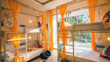 A dormitory room with bunk beds, orange curtains, and a view of trees through the windows.