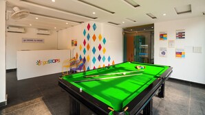 Une table de billard recouverte d’un drap de feutre vert, un mur blanc orné de motifs géométriques colorés, et une pancarte sur laquelle on peut lire : « Allez plus loin. Soyez plus. »