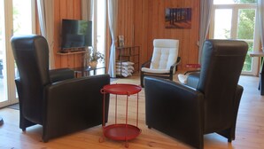 Una sala de estar con dos sillones de cuero negro, una butaca blanca, una mesita auxiliar roja, un televisor y un cuadro en la pared.