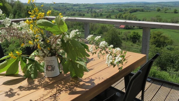Una terraza de madera con una mesa y sillas, una maceta con flores amarillas y blancas, y una vista panorámica de campos verdes y una casa a lo lejos.