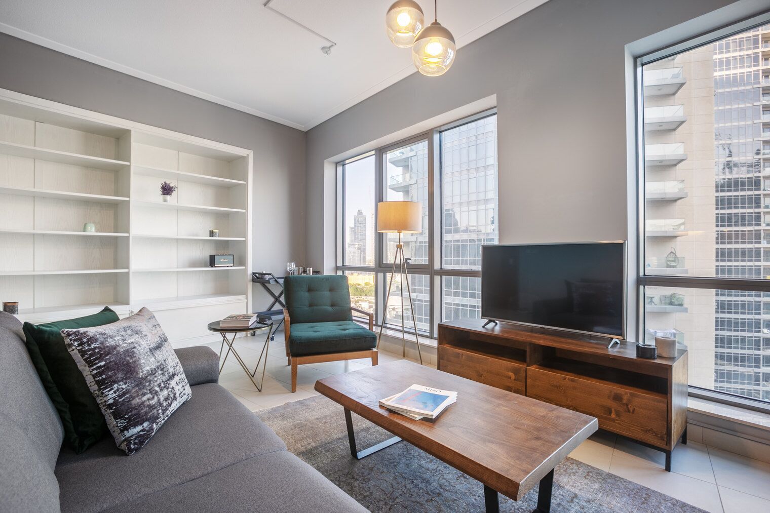 A modern living room with a sofa, armchair, coffee table, TV, and large windows offering a city view.
