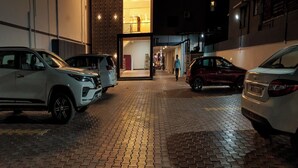 A modern building with a glass facade and a brick ground floor. There are parked cars on the street in front. - Grand Sri Krishna Residency (Viluppuram)