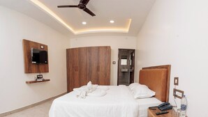 A modern bedroom with a curved bed, wooden headboard, a ceiling fan, a mounted TV, and a bedside table with a phone, glasses, and a water bottle. - Grand Sri Krishna Residency (Viluppuram)