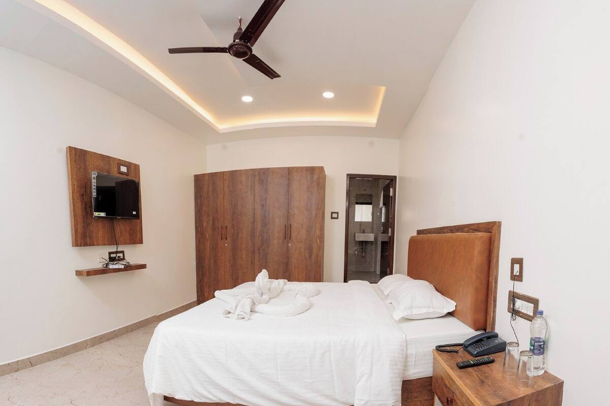 Een moderne slaapkamer met een gebogen bed, een houten hoofdbord, een plafondventilator, een wandgemonteerde televisie en een nachtkastje met een telefoon, bril en een waterfles.