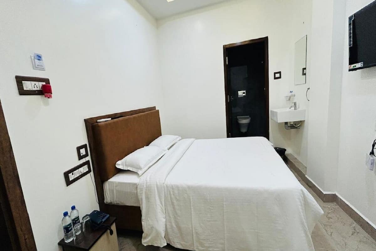 Een hotelkamer met een enkel bed, een nachtkastje met telefoon en waterflesjes, een klein kastje, een badkamer met wastafel en spiegel, en een deur die naar een andere kamer leidt.