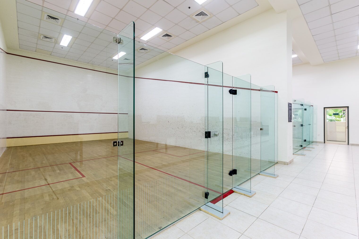 Una cancha de squash con paredes de vidrio y piso de madera, equipada con una puerta y un ventanal pequeño.