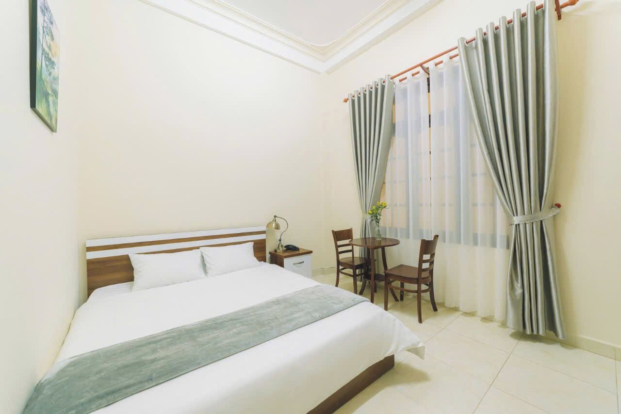Une chambre d’hôtel avec un lit, une table de chevet, une chaise, une petite table et une fenêtre avec des rideaux.