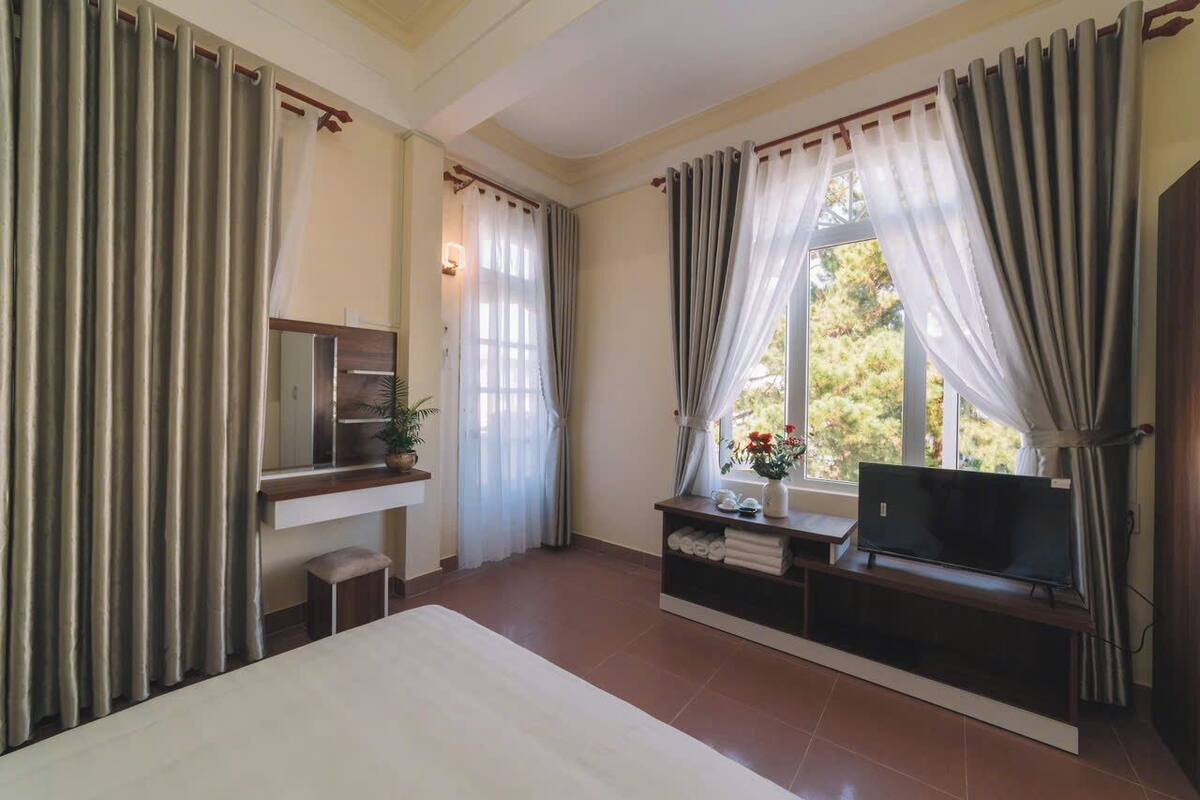Une chambre d’hôtel avec un lit, un téléviseur posé sur un meuble, un bureau avec un ordinateur et une fenêtre avec des rideaux.