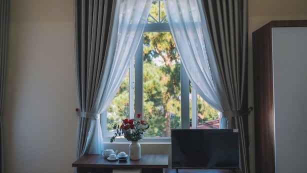 A room with a window, curtains, a small table with a vase of flowers, and a television. - Phat Loi Hotel Dalat  (Xuan Huong - Da Lat)