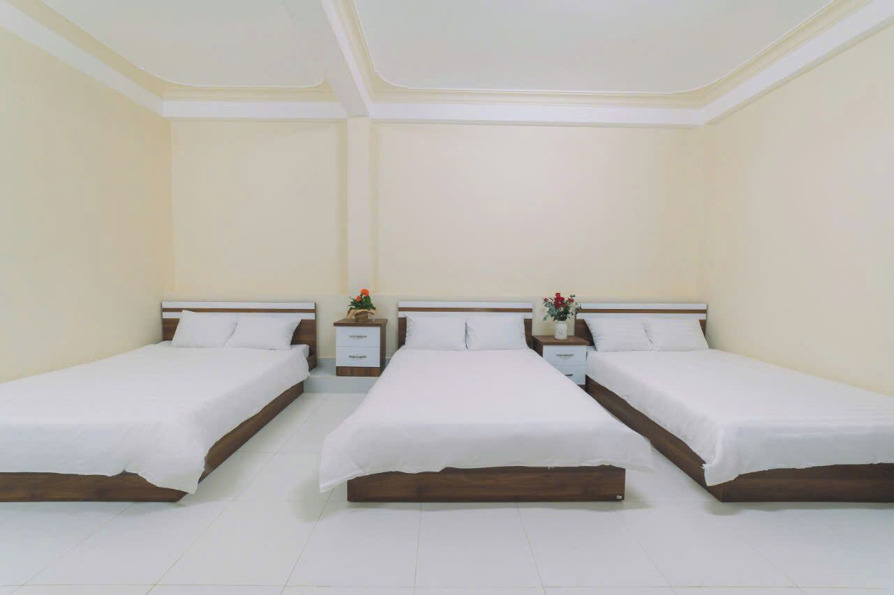 Habitación de hotel con tres camas individuales, ropa de cama blanca, cabeceros de madera y mesitas de noche con lámparas y pequeñas plantas.
