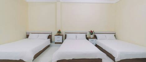 Habitación de hotel con tres camas individuales, ropa de cama blanca, cabeceros de madera y mesitas de noche con lámparas y pequeñas plantas.