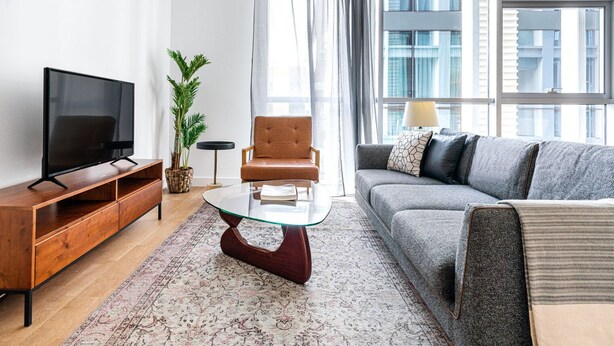 A modern living room with a flat-screen TV, a brown leather armchair, a grey sofa with patterned cushions, a glass coffee table, and a potted plant. - Blueground | City Walk, pool, nr City Park (Dubai)