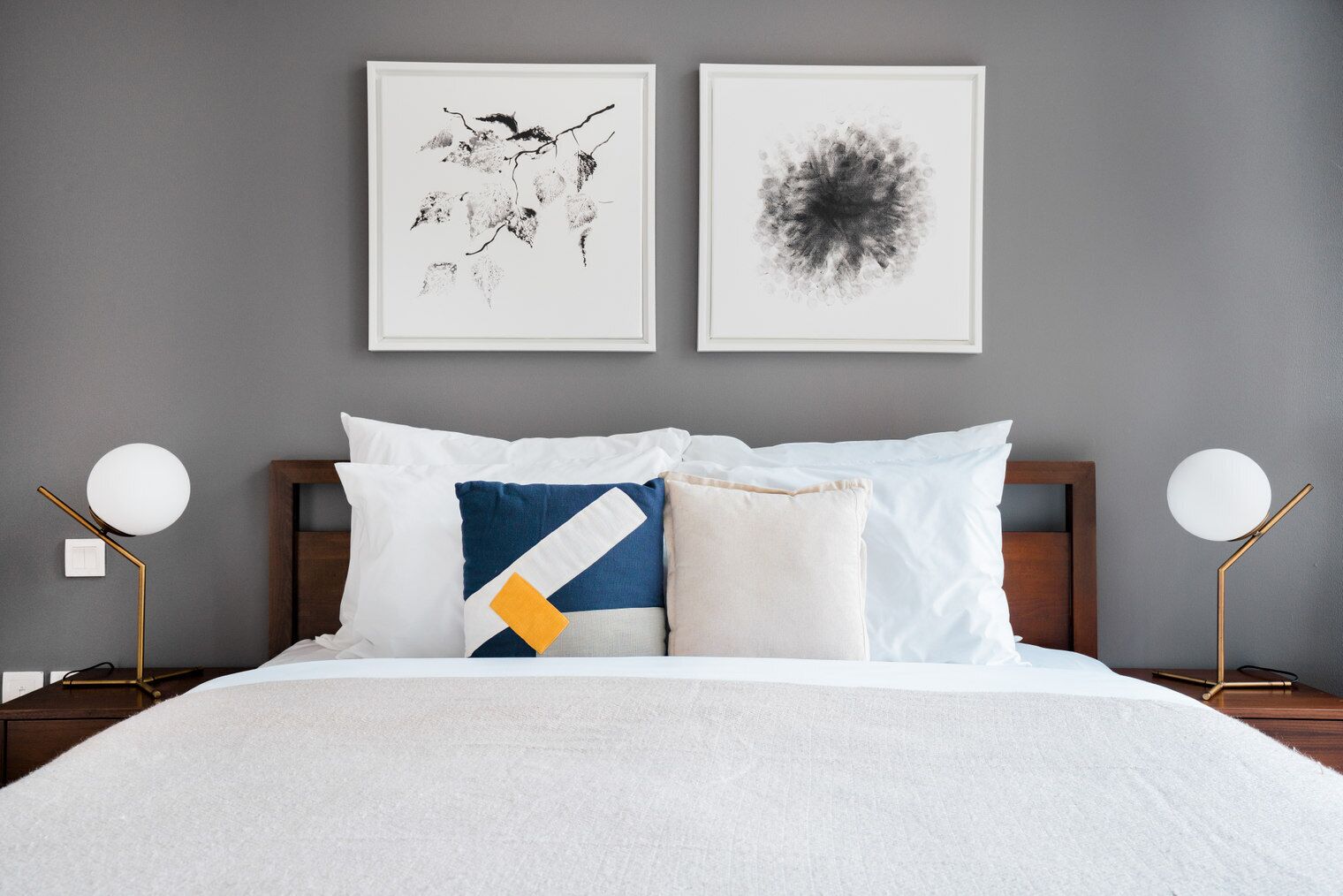 A neatly made bed with white bedding, a blue and yellow pillow, and two framed abstract artworks on the wall.