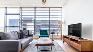 A modern living room with a grey sofa, teal armchair, wooden coffee table, and a TV on a wooden stand. Large windows offer a view of the cityscape.