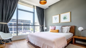 A modern bedroom with a large bed, a chair, a bedside table with a lamp, and two framed botanical prints on the wall.