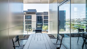 A modern balcony with a wooden table and chairs, large glass windows, and a view of surrounding buildings.