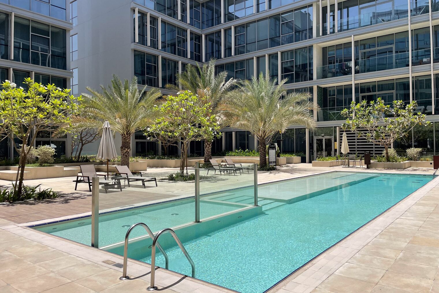 A modern hotel with a rectangular pool, lounge chairs, and palm trees.