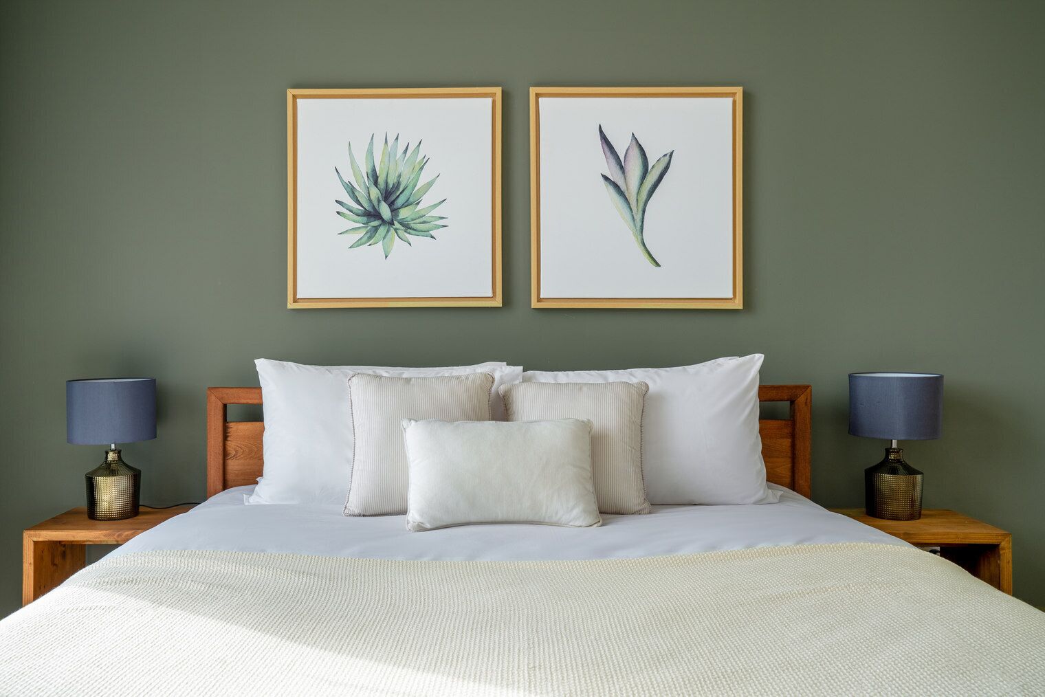 Una cama bien hecha con ropa blanca, dos láminas enmarcadas de motivos botánicos en la pared y dos lámparas de noche sobre mesitas de madera.
