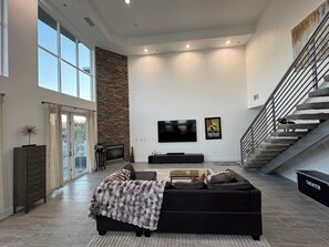 Relax in a spacious living area with a high ceiling and large windows, featuring a modern sofa, flat-screen TV, and decorative touches.
