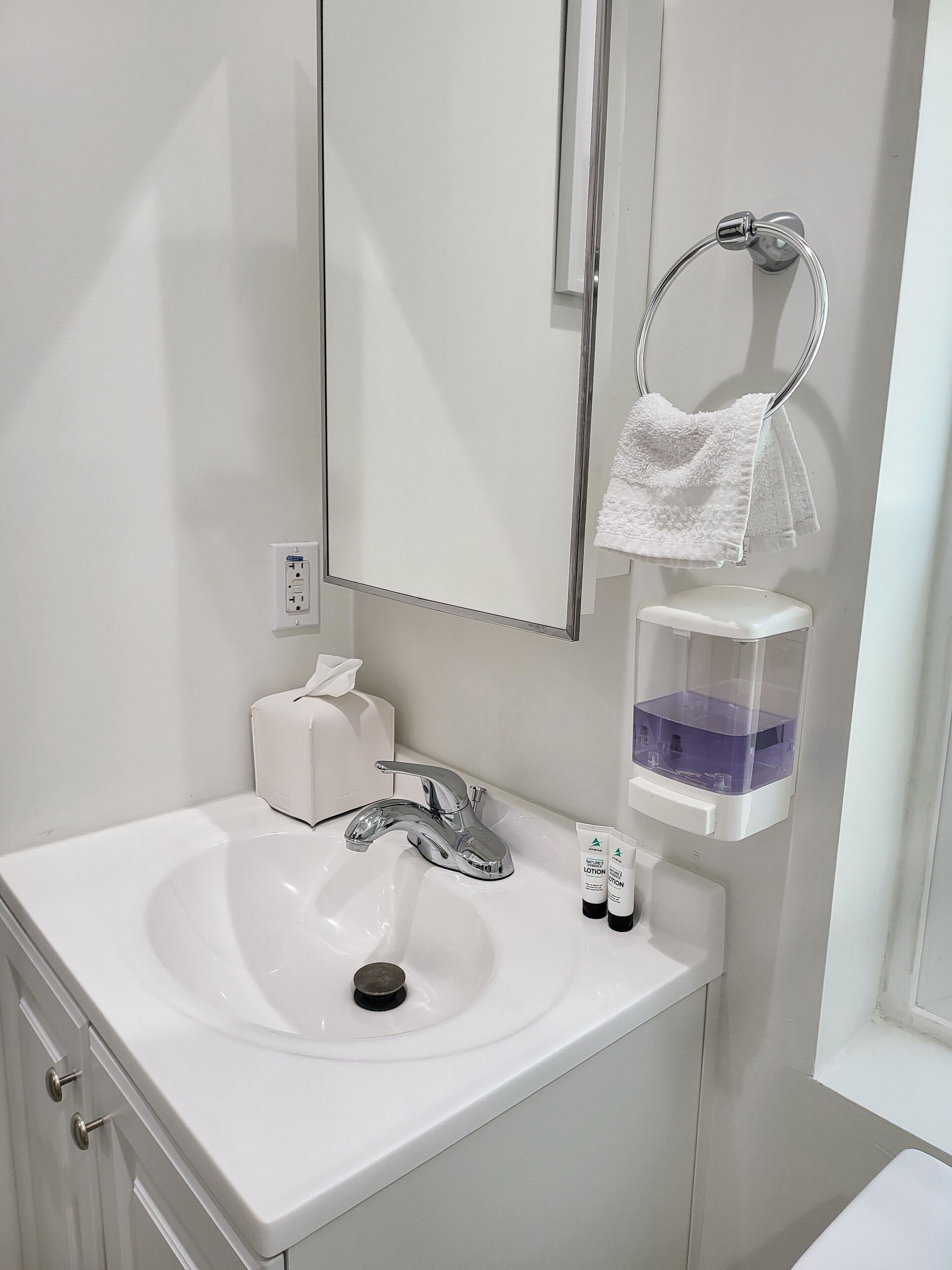 Un baño con un lavamanos blanco, un espejo, un perchero para toallas y un dispensador de jabón.