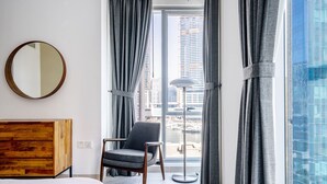 Moderni hotellihuone, jossa on suuri sänky, puinen yöpöytä, harmaa nojatuoli ja näkymä kaupunkimaisemaan suurista, lattiasta kattoon ulottuvista ikkunoista.