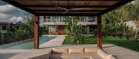 Un espacio de estar al aire libre techado, con piscina, palmeras y un edificio moderno al fondo.