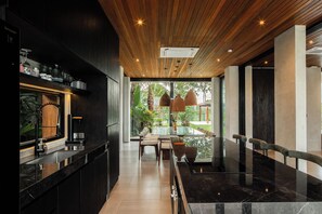 A modern interior with a wooden ceiling, a bar area, and a seating space with a view of greenery outside. - Famara Villa Bali by Blacksand  (Cepaka)