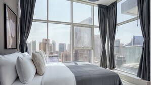 Una habitación de hotel moderna con una cama grande, cortinas grises y vistas a la ciudad a través de ventanas de ventanal.