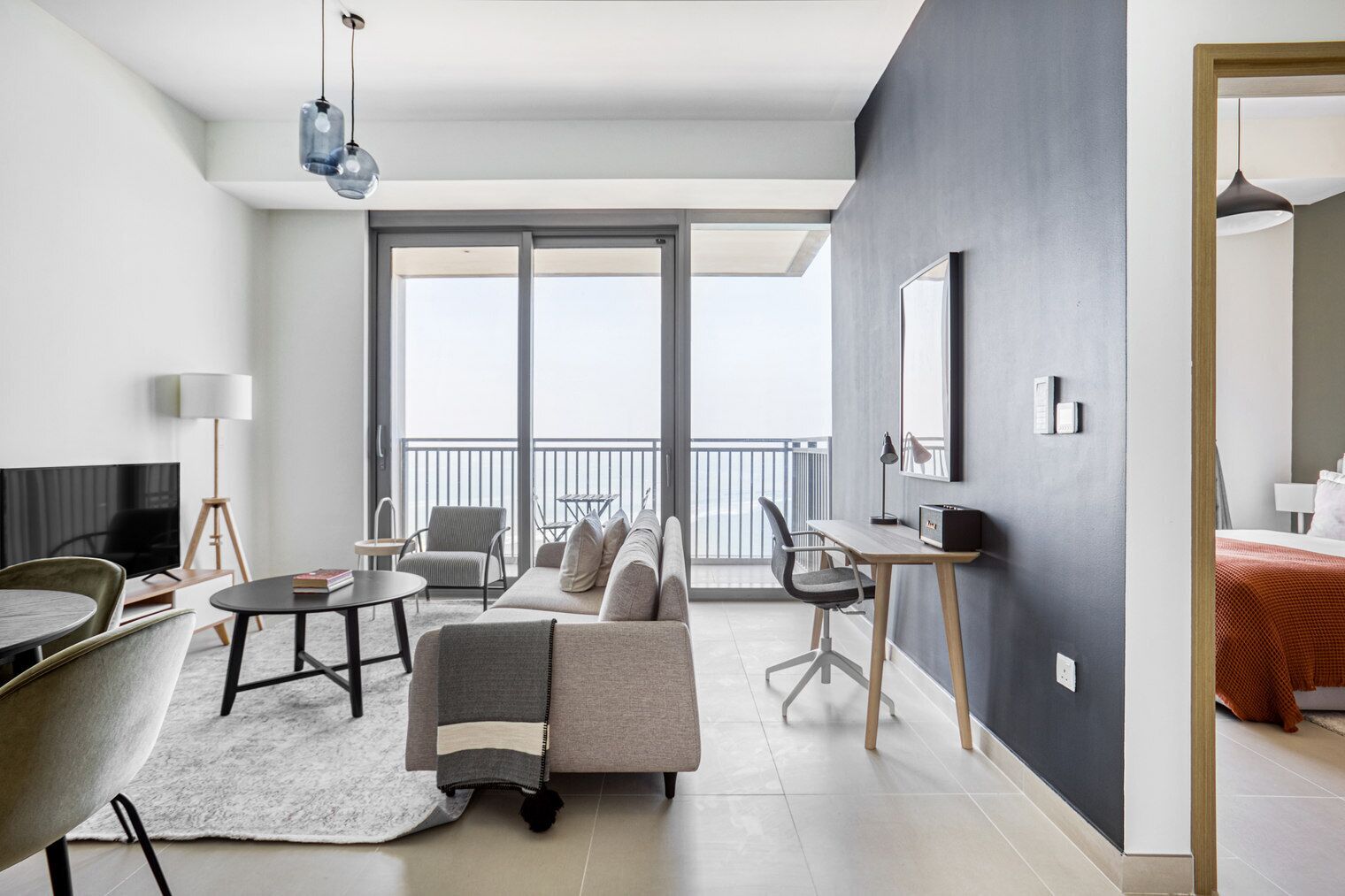 A modern living room with a sofa, coffee table, and TV. There is a balcony with a railing and a view of the sky.