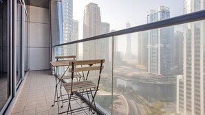 A balcony with two wooden chairs and a metal table, overlooking a cityscape with high-rise buildings and a river. - Blueground | JLT, pool, nr Marina (Dubai)