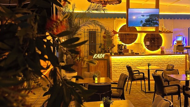 An indoor dining area with a long wooden table, chairs, hanging lights, a TV screen, and a bar area with a neon sign. - HANCI PANSİYON (Edremit)
