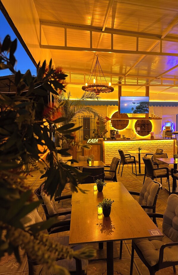 An indoor dining area with a long wooden table, chairs, hanging lights, a TV screen, and a bar area with a neon sign. - HANCI PANSİYON (Edremit)