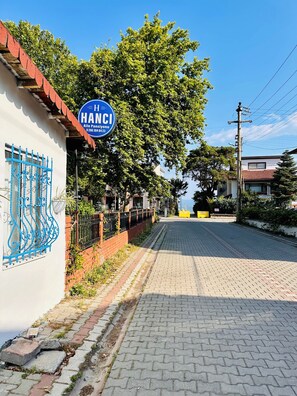 A street with a blue sign reading 'HANCI,' a paved road, and a building with a red roof. - HANCI PANSİYON (Edremit)