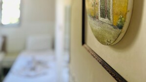 A framed artwork with a window scene, a bed, and a bedside table in the background. - HANCI PANSİYON (Edremit)