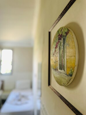 A framed artwork with a window scene, a bed, and a bedside table in the background. - HANCI PANSİYON (Edremit)