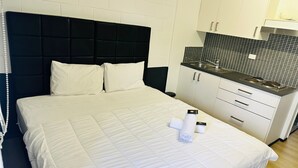A hotel room with a bed, a black headboard, white bedding, a small kitchen area with a sink and cabinets, and a tiled backsplash.
