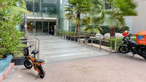 Un bâtiment moderne avec des fenêtres en verre, une entrée abritée et un espace pavé agrémenté de plantes en pot et de scooters garés.
