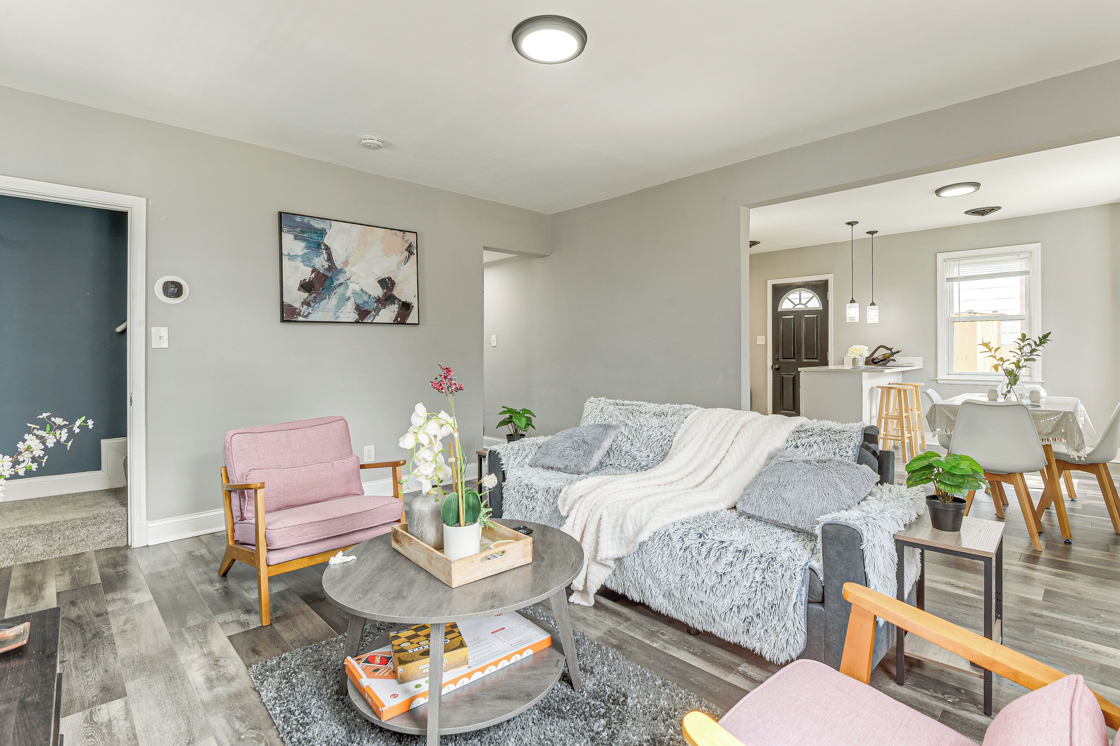 Ein modernes Wohnzimmer mit einem grauen Sofa, einem rosafarbenen Sessel und einem Couchtisch. An der Wand hängt ein Gemälde und auf dem Tisch steht eine Pflanze.