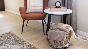 A room with a modern chair, a small round table with a clock, a patterned rug, and a window with curtains. - Thankers (Wheatland)