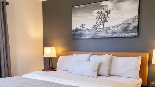 A neatly made bed with white linens and a gray throw blanket, a large abstract painting on the wall, and two bedside lamps. - Thankers (Wheatland)