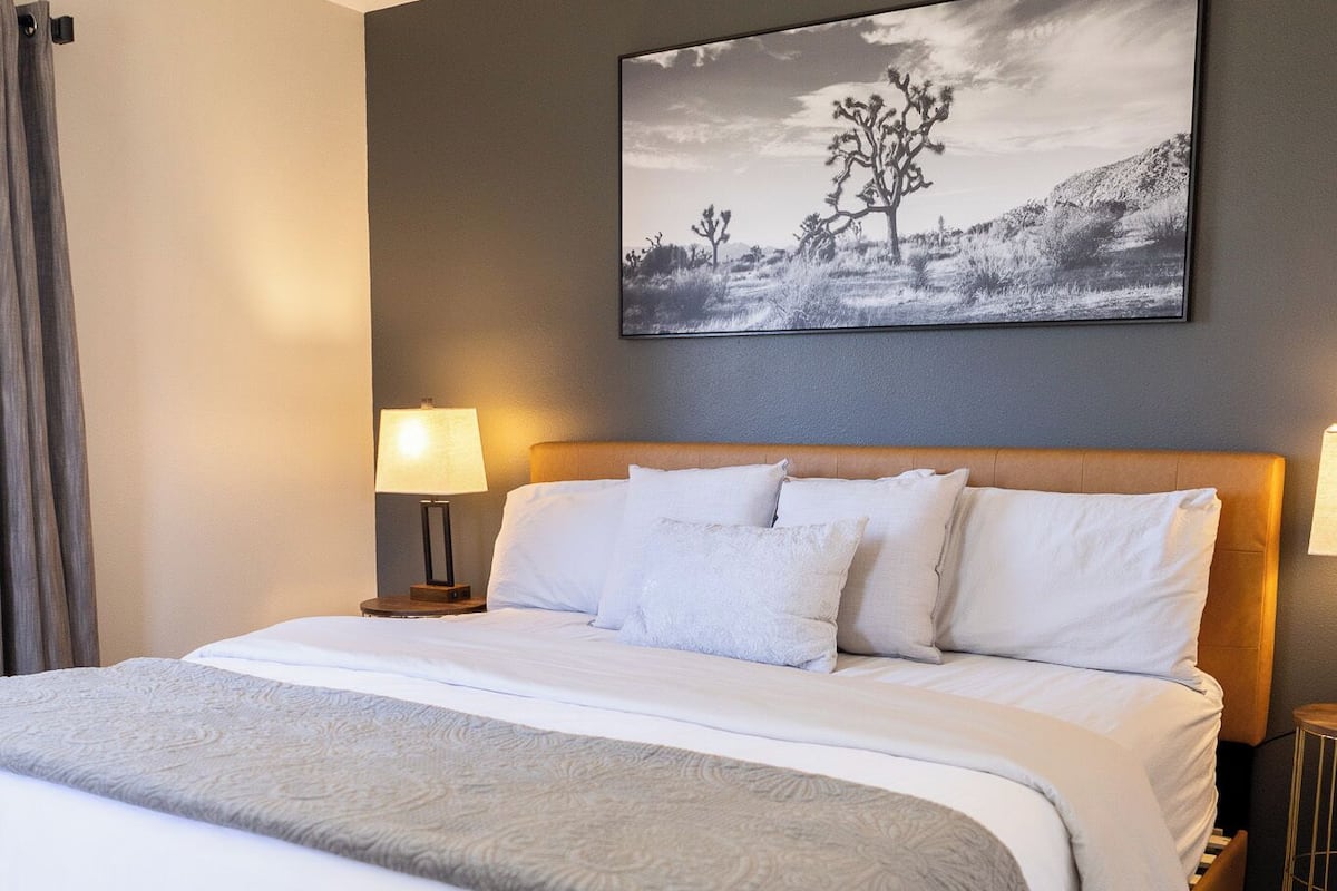 A neatly made bed with white linens and a gray throw blanket, a large abstract painting on the wall, and two bedside lamps.