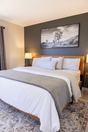 A neatly made bed with white linens and a gray throw blanket, a large abstract painting on the wall, and two bedside lamps. - Thankers (Wheatland)
