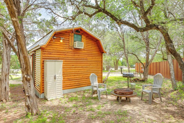 A wooden cabin with a small porch, two Adirondack chairs, and a round table. - Cozy Couple's Cabin Retreat in Concan! (Concan)