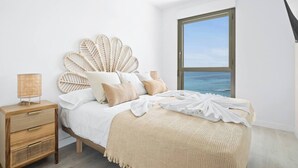 Ein Schlafzimmer mit großem Fenster, einem Bett mit Blattranken-Verziertem Kopfteil, einem Nachttisch mit Lampe und Blick aufs Meer.