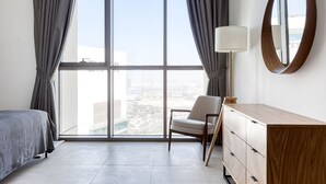 A modern hotel room with a large window, a bed, a chair, a dresser, and a round mirror.