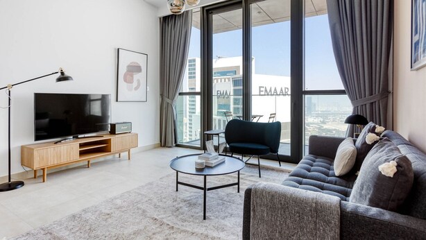 A modern living room with a large flat-screen TV, a sofa, a coffee table, and a view of the cityscape through large windows.
