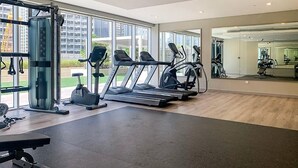 A modern gym with cardio machines, weight equipment, and large windows offering views of the city.